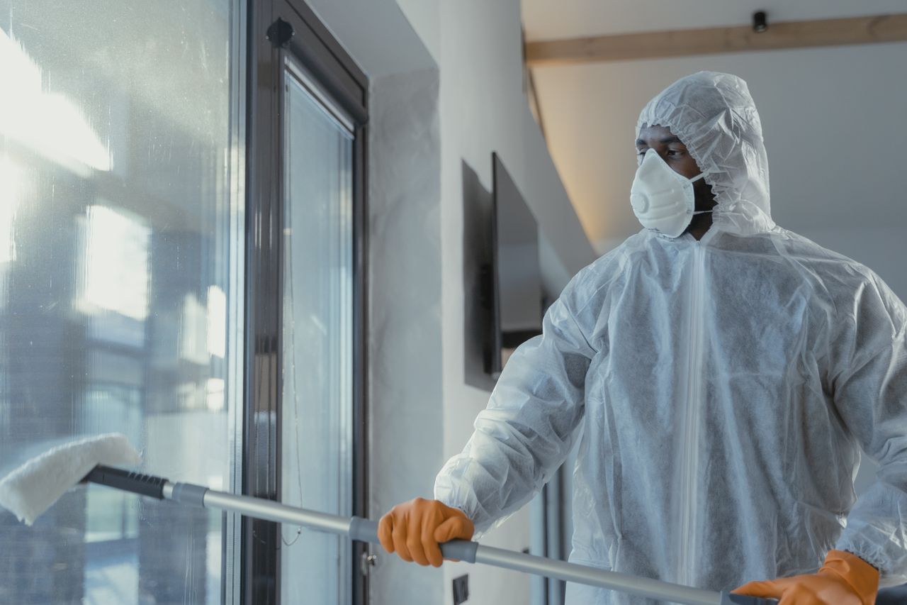 An adult male cleaner wearing protective gear and a mask is washing a window indoors on a bright sunny day.